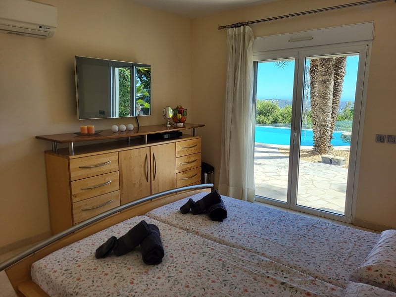 Schlafzimmer mit zwei Einzelbetten, Wandfernseher über einer Holzkommode und direktem Zugang zur Außenterrasse mit Pool und Palmen.