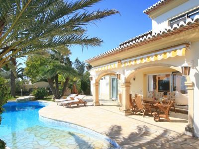 Der Außenbereich der Villa zeigt einen Freiform-Swimmingpool und eine weitläufige Steinterrasse, die zu einer überdachten Naya mit Holz-Essgruppe und gestreiften Markisen führt.