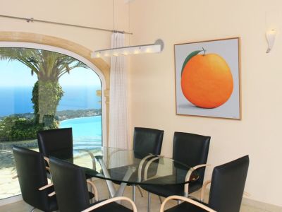 Essbereich mit Glastisch und modernen Stühlen, akzentuiert durch ein großes Rundbogenfenster mit Panoramablick auf Meer und Küste. Ein Kunstwerk mit einer Orange ziert die Wand.