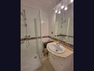 Funktionales Badezimmer mit begehbarer Glasdusche, Waschtisch mit hellem Marmor-Arbeitsplatte und weißem Unterschrank. Weiße Wandfliesen sind mit einem dekorativen mediterranen Bordürenband versehen.