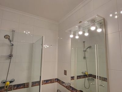 Funktionales Badezimmer mit weißen Fliesen und dekorativer Bordüre, Glasduschkabine und Waschtisch mit Marmor-Effekt-Arbeitsplatte. Beleuchteter Spiegel und blaue Deckenleuchte.