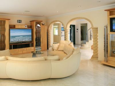 Geräumiges Wohnzimmer mit großem, geschwungenem Cremesofa, hellem Marmorboden und maßgefertigten hellen Holzmöbeln. Steinerne Bögen mit Säulen führen zum Eingangsbereich.