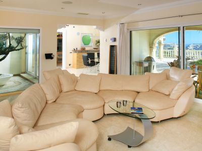 Geräumiges Wohnzimmer mit hellem Ecksofa, Glastisch und Marmorboden. Große Fensterfronten öffnen sich zu einer Meerblickterrasse und einem Innenhof mit Olivenbaum.
