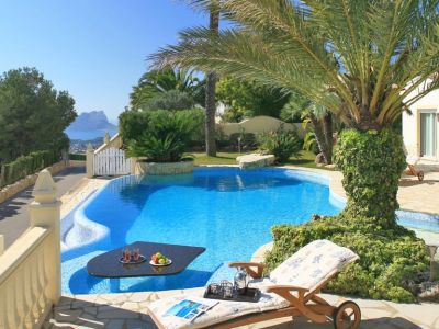 Geschwungener Pool mit einem versenkten schwarzen Tisch und Erfrischungen, angrenzend an eine steingepflasterte Terrasse mit Sonnenliege. Ferne Meerblicke und eine markante Felsformation sind sichtbar.