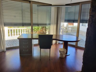 Großzügiges privates Büro mit maßgeschneidertem Holzschreibtisch und raumhohen Fenstern, die den Blick auf eine Kiesterrasse und mediterrane Pflanzen freigeben. Dunkler Parkettboden.