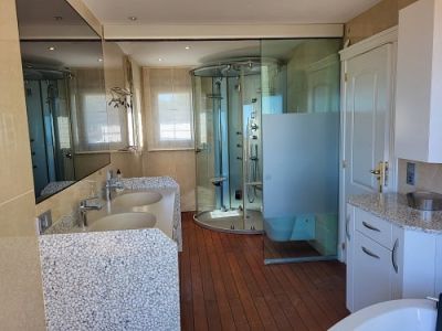 Hauptbadezimmer mit Doppelwaschbecken auf einzigartiger Kieselsteinbasis, geräumiger Glas-Massagedusche und warmem Holzfußboden. Natürliches Licht durch Fenster.