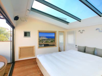 Helles Schlafzimmer mit großem Glasdachfenster und warmem Holzboden. Eine Schiebetür führt zur privaten Terrasse mit Korbsessel; ein eingebauter TV zeigt Küstenblicke.