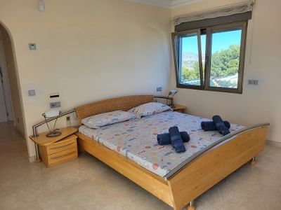 Helles Schlafzimmer mit hellem Holzbett, geflochtenem Kopfteil und Nachttisch. Das offene Fenster bietet Ausblicke auf üppiges Grün und ferne Berge.