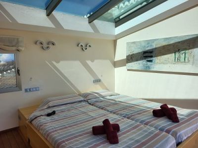 Helles Schlafzimmer mit zwei Einzelbetten, gestreifter Bettwäsche und roten Handtüchern. Ein großes Dachfenster sorgt für natürliches Licht, ergänzt durch ein abstraktes Gemälde an der Wand.