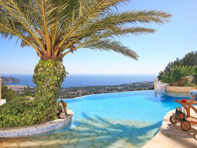 Infinity-Pool mit Panoramablick auf Meer und Küste, inklusive einer von Efeu bewachsenen Palme und einem Holz-Servierwagen auf der Steinterrasse.