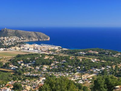 Panoramablick von einem Hügel, mit einer Küstenstadt, einem Yachthafen, einem markanten Berg und dem tiefblauen Mittelmeer unter klarem Himmel.