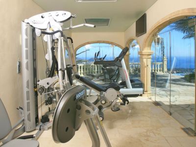 Privater Fitnessraum mit Hoist- und Life Fitness-Geräten, Marmorboden und gewölbten Glaswänden mit Panoramablick auf Meer und Pool. Dachfenster sorgen für Tageslicht.