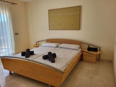 Schlafzimmer mit Holzbettrahmen, geflochtenem Kopfteil und Metallakzenten. Nachttische mit Schubladen flankieren das Bett unter einem abstrakten Gemälde. Tageslicht fällt durch transparente Vorhänge.