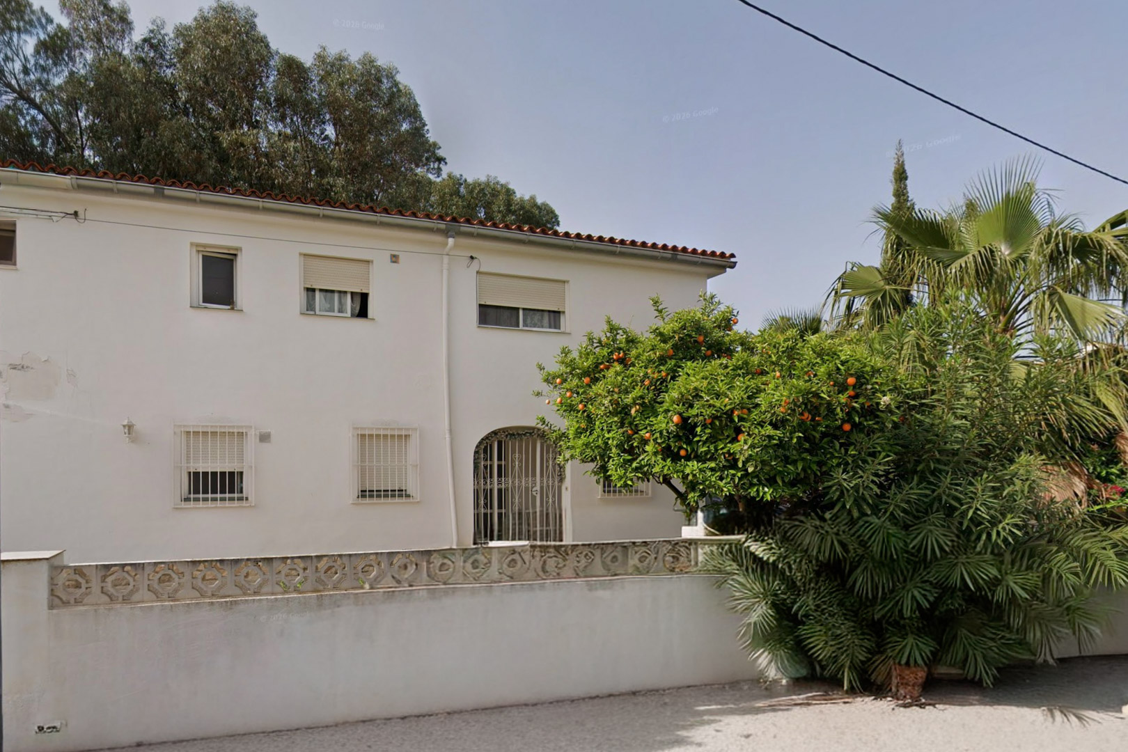 Außenansicht der weißen Villa mit rotem Ziegeldach, vergitterten Fenstern und einem bogenförmigen Eingang. Ein Orangenbaum mit Früchten und üppige Vegetation prägen den Vorgarten.