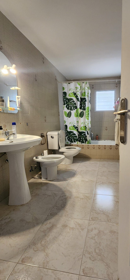 Badezimmer mit hellen Fliesen, Standwaschbecken, Toilette, Bidet und Badewanne mit tropischem Duschvorhang sowie einem Milchglasfenster.
