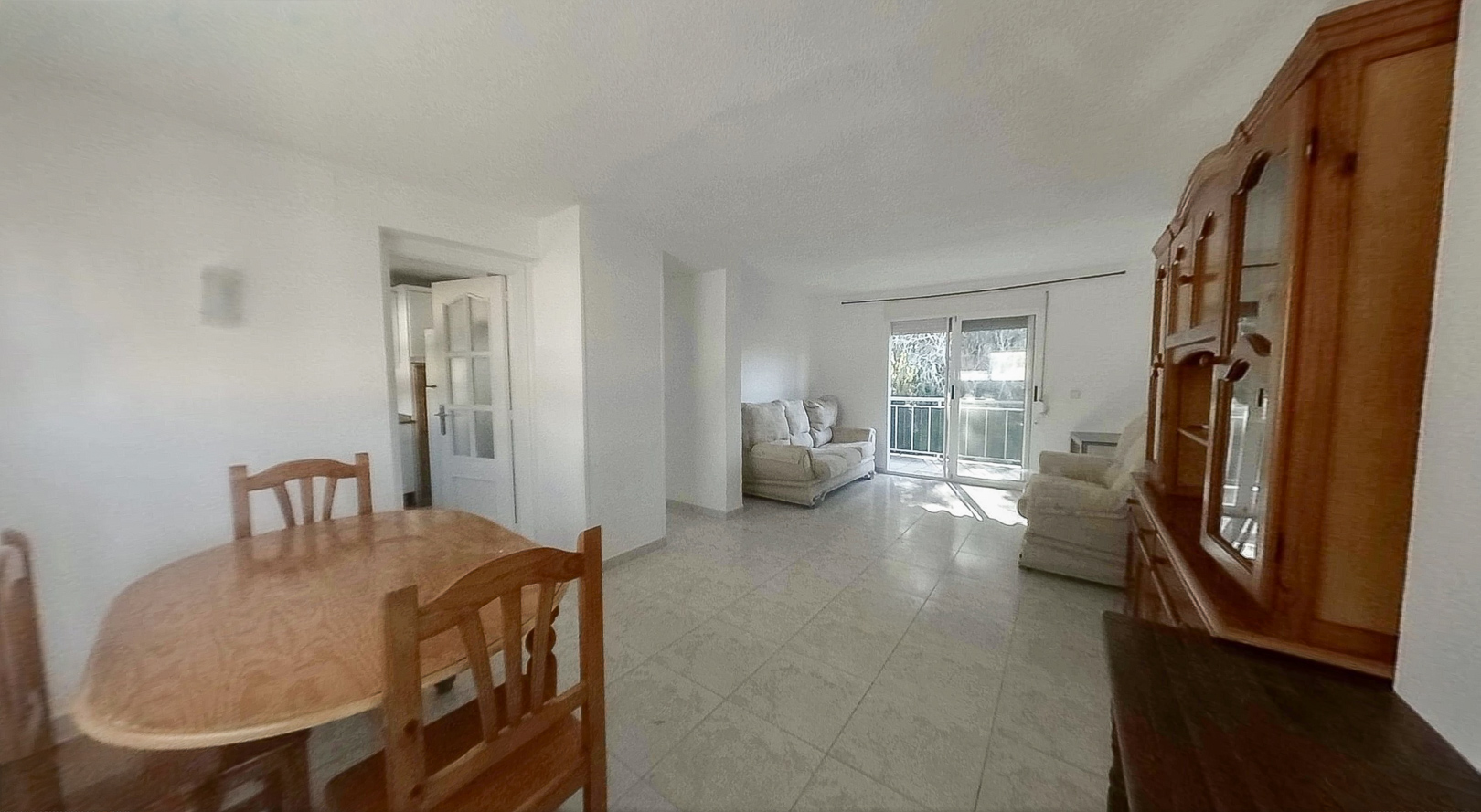 Blick auf den hellen Wohn-Essbereich mit ovalem Holztisch, Fliesenboden und großem Holzschrank. Glasschiebetüren führen zu einem Balkon und durchfluten den Raum mit natürlichem Licht.