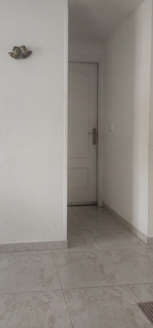 Blick auf einen hellen Flur mit hellen Fliesenböden und weißen strukturierten Wänden. Eine dekorative Puttenverzierung schmückt die linke Wand und führt zu einer weißen Tür mit silbernem Griff.