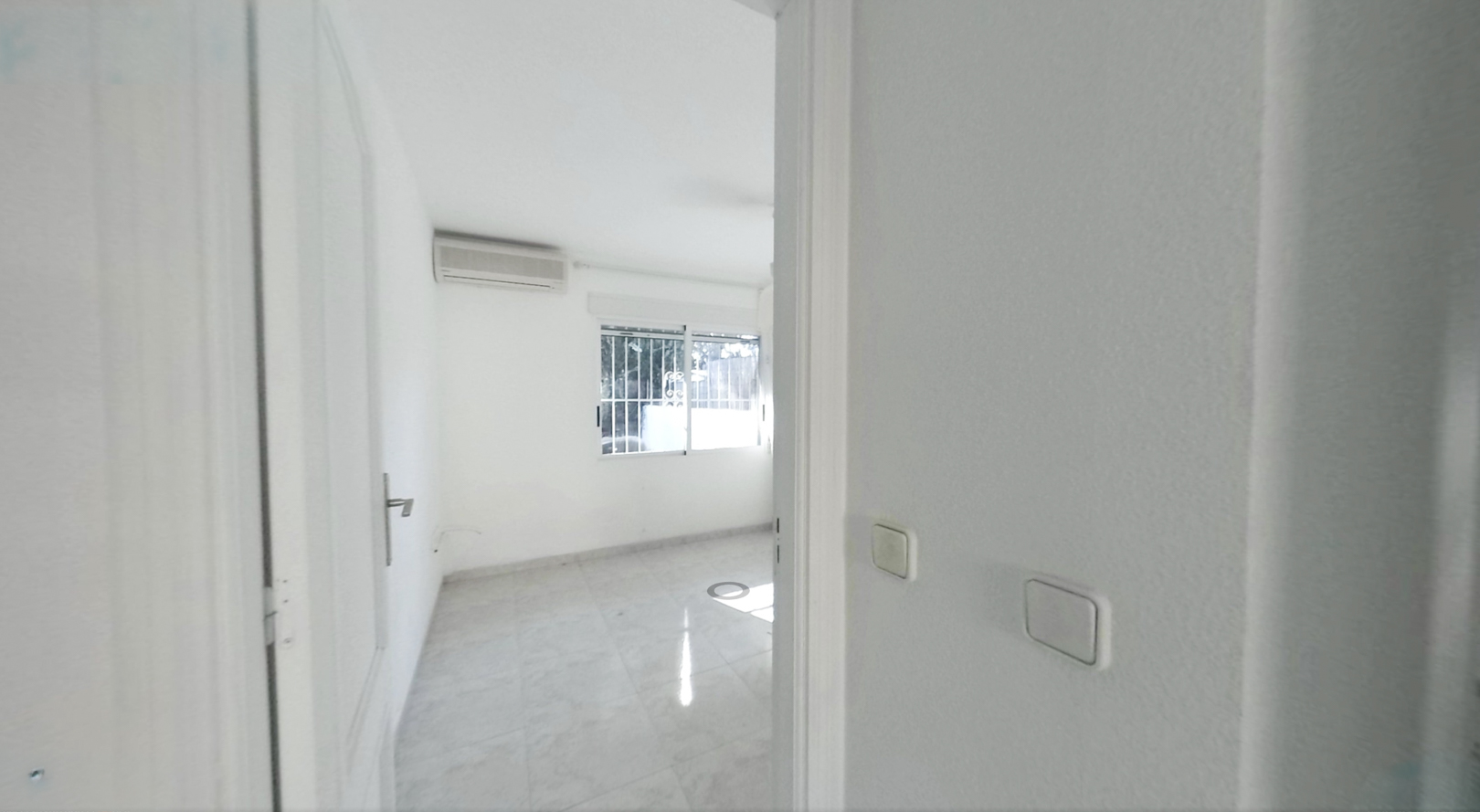 Blick in einen leeren Raum mit hellen Fliesenböden, weißen Wänden und einer Klimaanlage. Das Fenster mit Sicherheitsgittern lässt Tageslicht herein.
