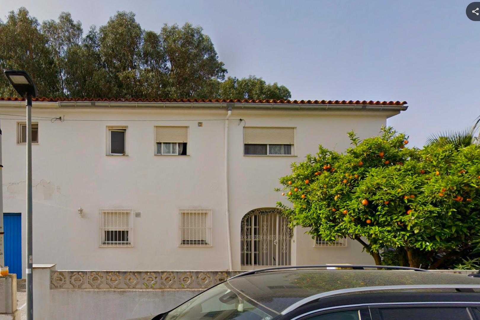 Die weiße Fassade der zweistöckigen Villa zeigt ein Ziegeldach, einen gewölbten Eingang mit Ziergitter und Fenster mit Gittern. Ein reifer Orangenbaum, voll mit Früchten, steht prominent davor.