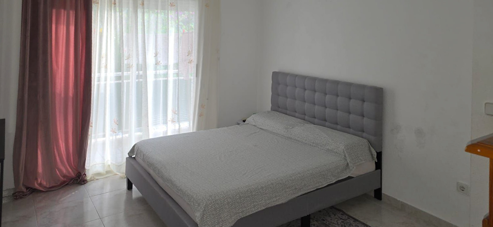 Doppelzimmer mit grauem Polsterbett und gestepptem Kopfteil, Fliesenboden und viel Tageslicht durch das Fenster mit Zugang zur Terrasse.