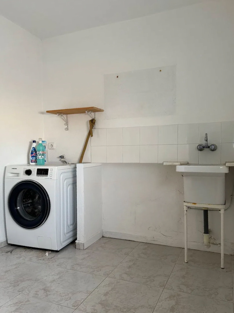 Funktionaler Waschraum mit Waschmaschine, praktischem Spülbecken und weiß gefliestem Spritzschutz. Heller Fliesenboden und ein kleines Holzregal sind sichtbar.