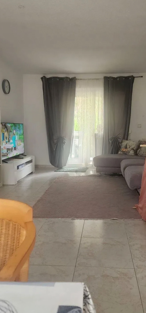Geräumiges Wohnzimmer mit hellen Fliesen, einem weichen, neutralen Teppich und einem bequemen grauen Modulsofa. Natürliches Licht fällt durch die Glastür mit Vorhängen, die zur Terrasse führt.