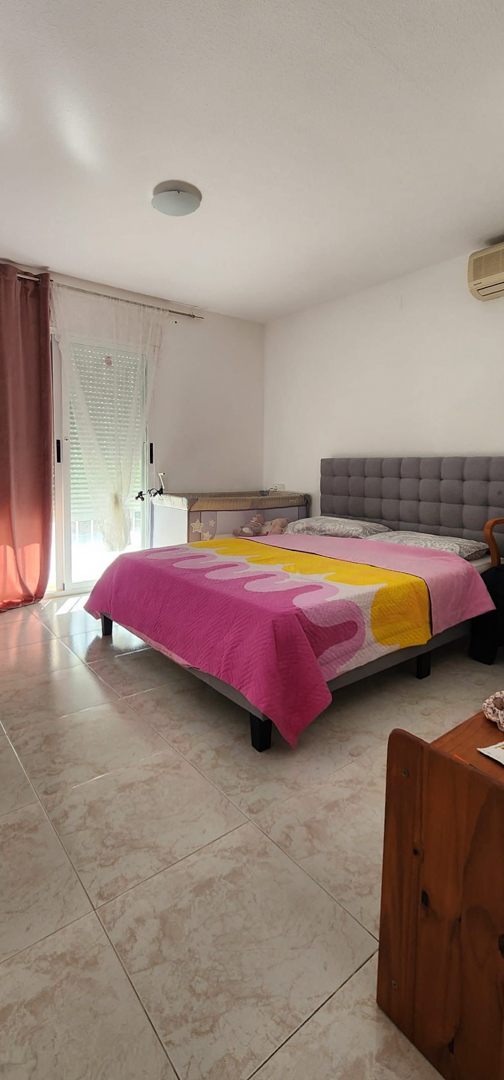 Helles Schlafzimmer mit Doppelbett, gepolstertem Kopfteil und Fliesenboden. Direkter Zugang nach draußen und Klimaanlage vorhanden.