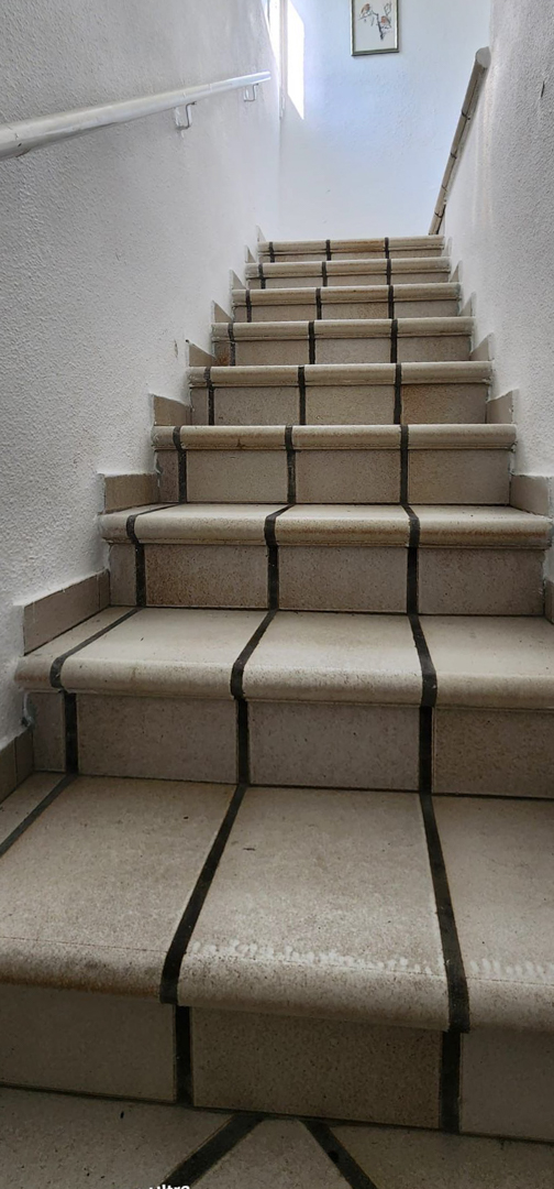 Innentreppe mit hellen Fliesenstufen und dunklen Fugen, strukturierten weißen Wänden und schlichten Handläufen. Tageslicht erhellt den oberen Bereich.