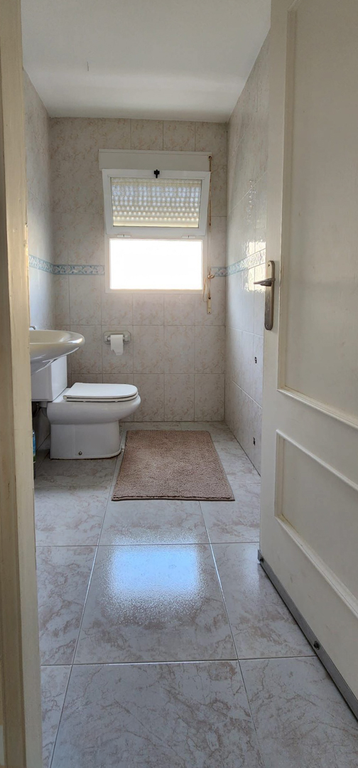 Kompaktes Badezimmer mit WC, wandmontiertem Waschbecken und Fenster mit Jalousie. Helle Fliesen an Wänden und Boden.