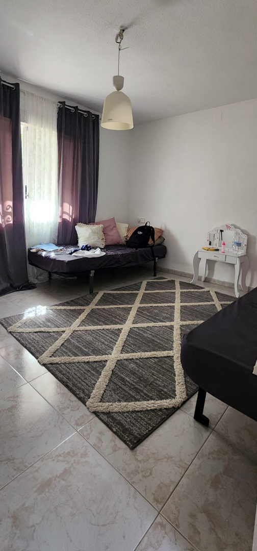 Schlafzimmer mit hellen Fliesen, einem grauen geometrischen Teppich und einem Tagesbett. Ein Fenster mit dunklen Vorhängen und ein weißer Schminktisch sind vorhanden.