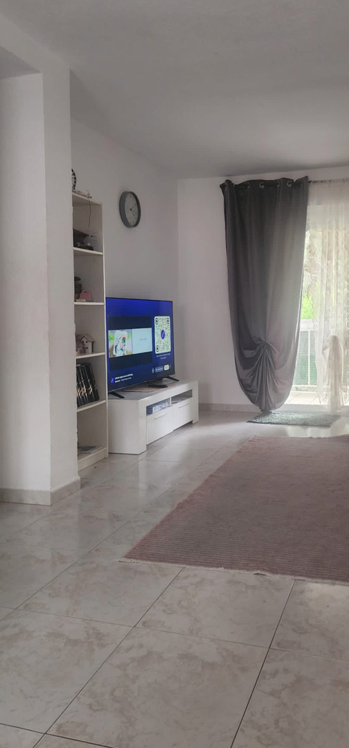 Teilansicht des Wohnzimmers mit Fernseher auf weißem Schrank, Regal und Zugang zur Terrasse mit grauen und transparenten Vorhängen, auf hellen Fliesenboden mit Teppich.