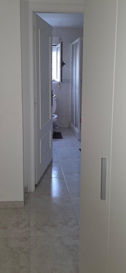 Teilansicht eines Badezimmers mit hellem Fliesenboden, Toilette, Fenster für Tageslicht und Duschkabine. Die Wandfliesen zeigen eine dekorative Bordüre.