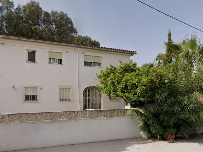 Außenansicht der weißen Villa mit rotem Ziegeldach, vergitterten Fenstern und einem bogenförmigen Eingang. Ein Orangenbaum mit Früchten und üppige Vegetation prägen den Vorgarten.