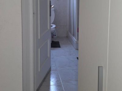 Blick in ein Badezimmer mit hellen Fliesen auf dem Boden und weißen Wandfliesen mit Zierbordüre. Es sind eine Toilette, eine Duschkabine und ein Fenster sichtbar.