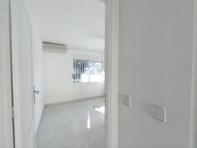 Blick in einen leeren Raum mit hellen Fliesenböden, weißen Wänden und einer Klimaanlage. Das Fenster mit Sicherheitsgittern lässt Tageslicht herein.
