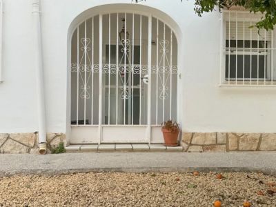 Der Haupteingang der Villa zeigt ein sicheres, bogenförmiges Metalltor. Ein Kiesgarten mit herabgefallenen Früchten und einem überhängenden Baum umrahmt die Fassade.