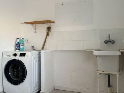 Funktionaler Waschraum mit Waschmaschine, praktischem Spülbecken und weiß gefliestem Spritzschutz. Heller Fliesenboden und ein kleines Holzregal sind sichtbar.