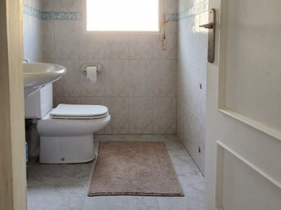 Funktionales Badezimmer mit Toilette, Waschbecken und Fenster. Es verfügt über hellcremefarbene Wand- und Bodenfliesen mit blauer Dekorleiste und eine Badematte.