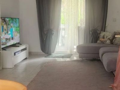 Geräumiges Wohnzimmer mit hellen Fliesen, einem weichen, neutralen Teppich und einem bequemen grauen Modulsofa. Natürliches Licht fällt durch die Glastür mit Vorhängen, die zur Terrasse führt.