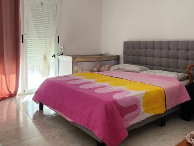 Helles Schlafzimmer mit Doppelbett, gepolstertem Kopfteil und Fliesenboden. Direkter Zugang nach draußen und Klimaanlage vorhanden.