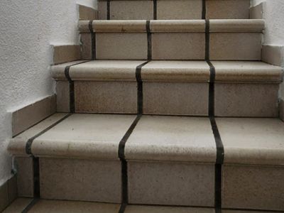 Innentreppe mit hellen Fliesenstufen und dunklen Fugen, strukturierten weißen Wänden und schlichten Handläufen. Tageslicht erhellt den oberen Bereich.