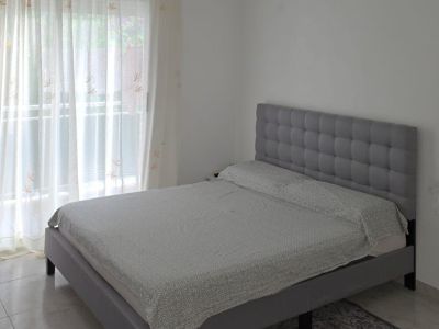 Schlafzimmer mit grauem Polsterbett und gestepptem Kopfteil. Tageslicht fällt durch das große Fenster mit transparenten und weinroten Vorhängen auf den Fliesenboden.
