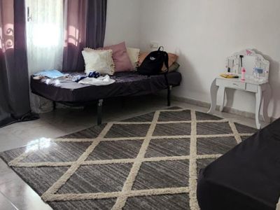 Schlafzimmer mit hellen Fliesen, einem geometrischen grau-beigen Teppich und einem Einzelbett am Fenster mit dunklen Vorhängen. Ein kleiner weißer Schminktisch ist sichtbar.
