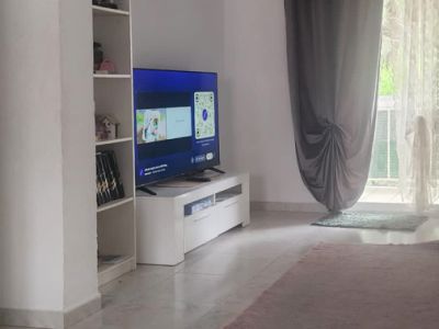 Teilansicht des Wohnzimmers mit Fernseher auf weißem Schrank, Regal und Zugang zur Terrasse mit grauen und transparenten Vorhängen, auf hellen Fliesenboden mit Teppich.