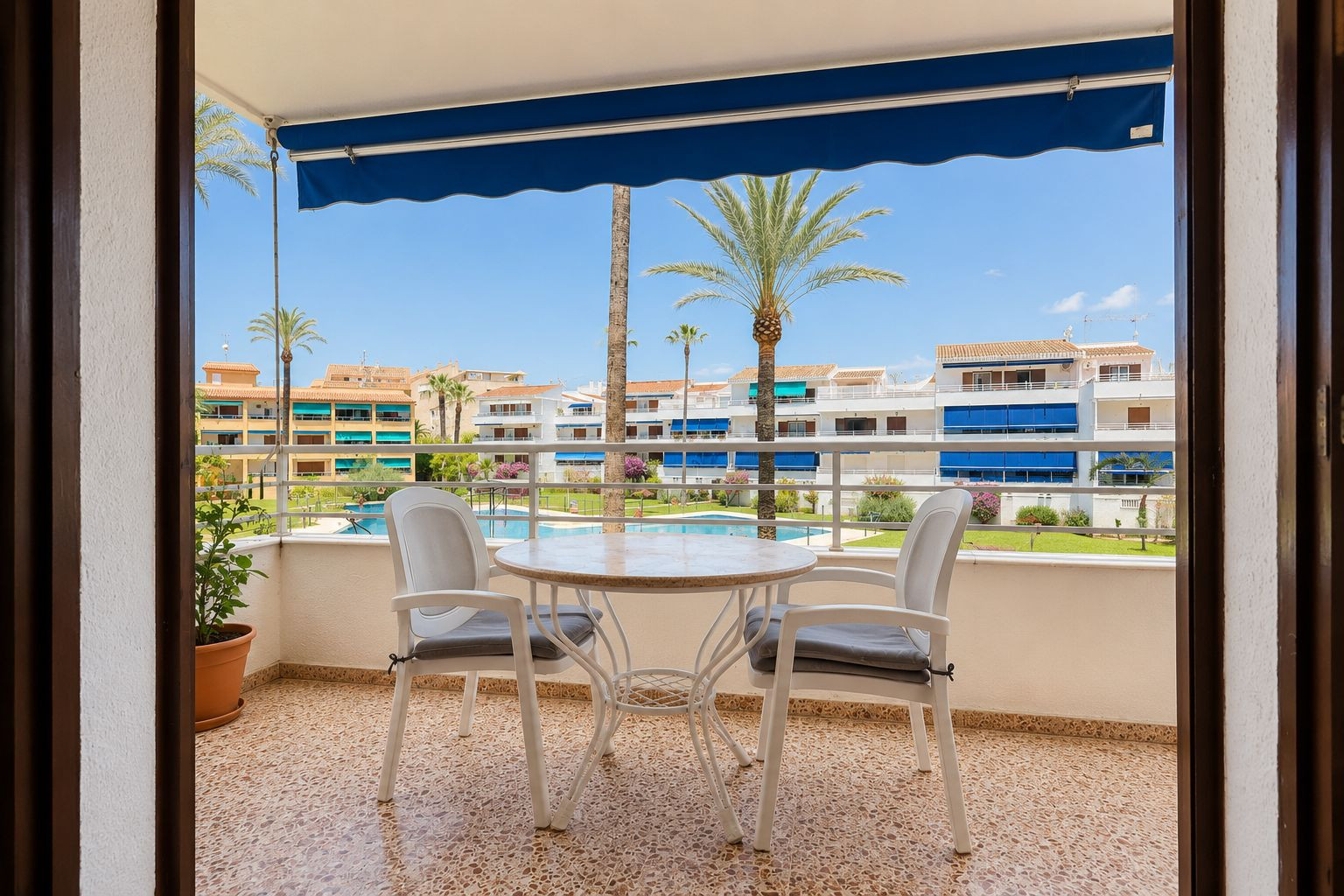 Geflieste Terrasse mit einem runden Tisch und zwei Stühlen, beschattet von einer blauen Markise. Direkter Blick auf den Gemeinschaftspool und Palmen. Geflieste Terrasse mit einem runden Tisch und zwei Stühlen, beschattet von einer blauen Markise. Direkter Blick auf den Gemeinschaftspool und Palmen.