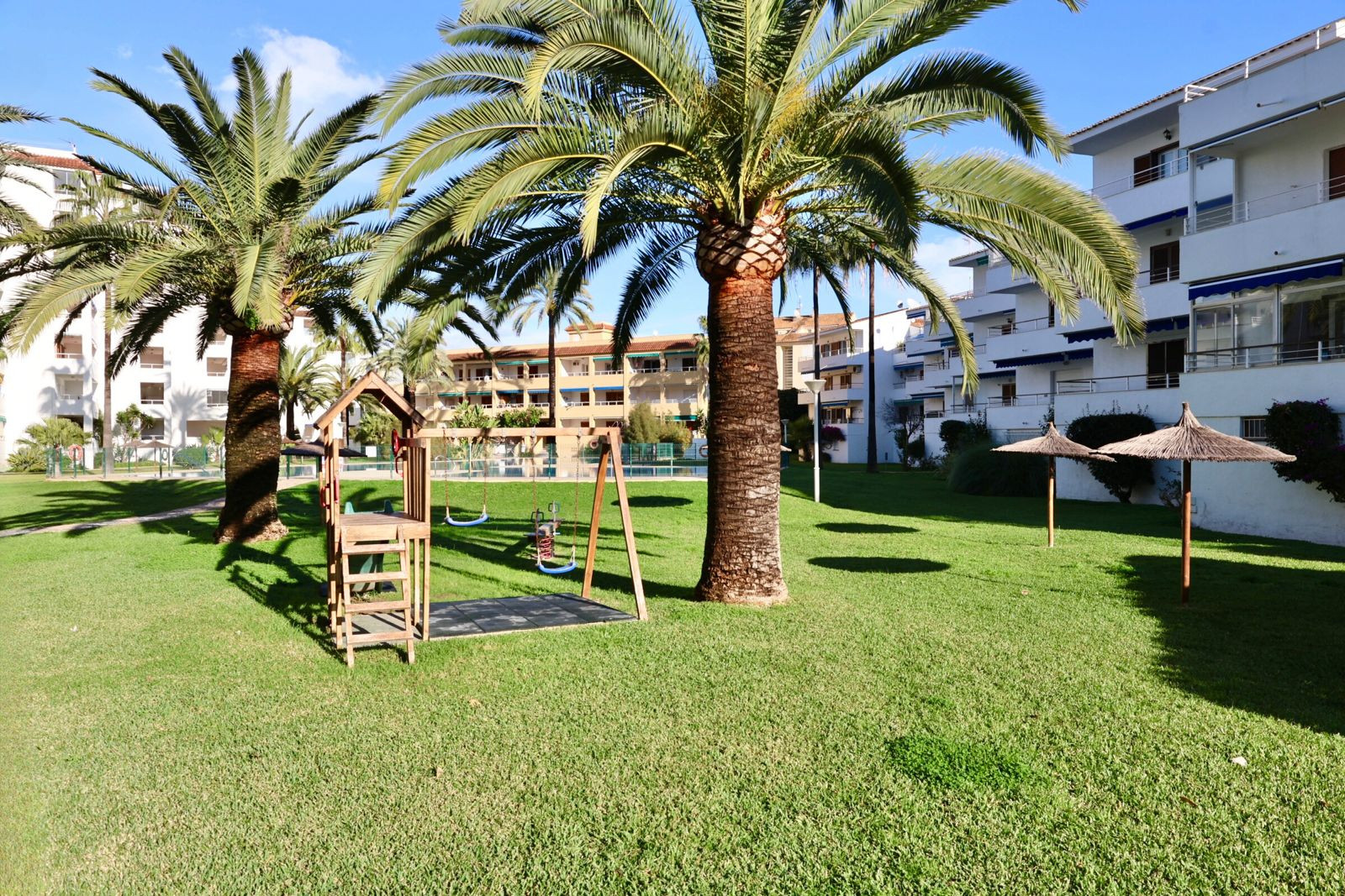 Gepflegte Gemeinschaftsgartenanlage mit Rasen, Holzspielplatz, hohen Palmen und Strohschirmen, umgeben von Wohngebäuden unter blauem Himmel. Gepflegte Gemeinschaftsgartenanlage mit Rasen, Holzspielplatz, hohen Palmen und Strohschirmen, umgeben von Wohngebäuden unter blauem Himmel.