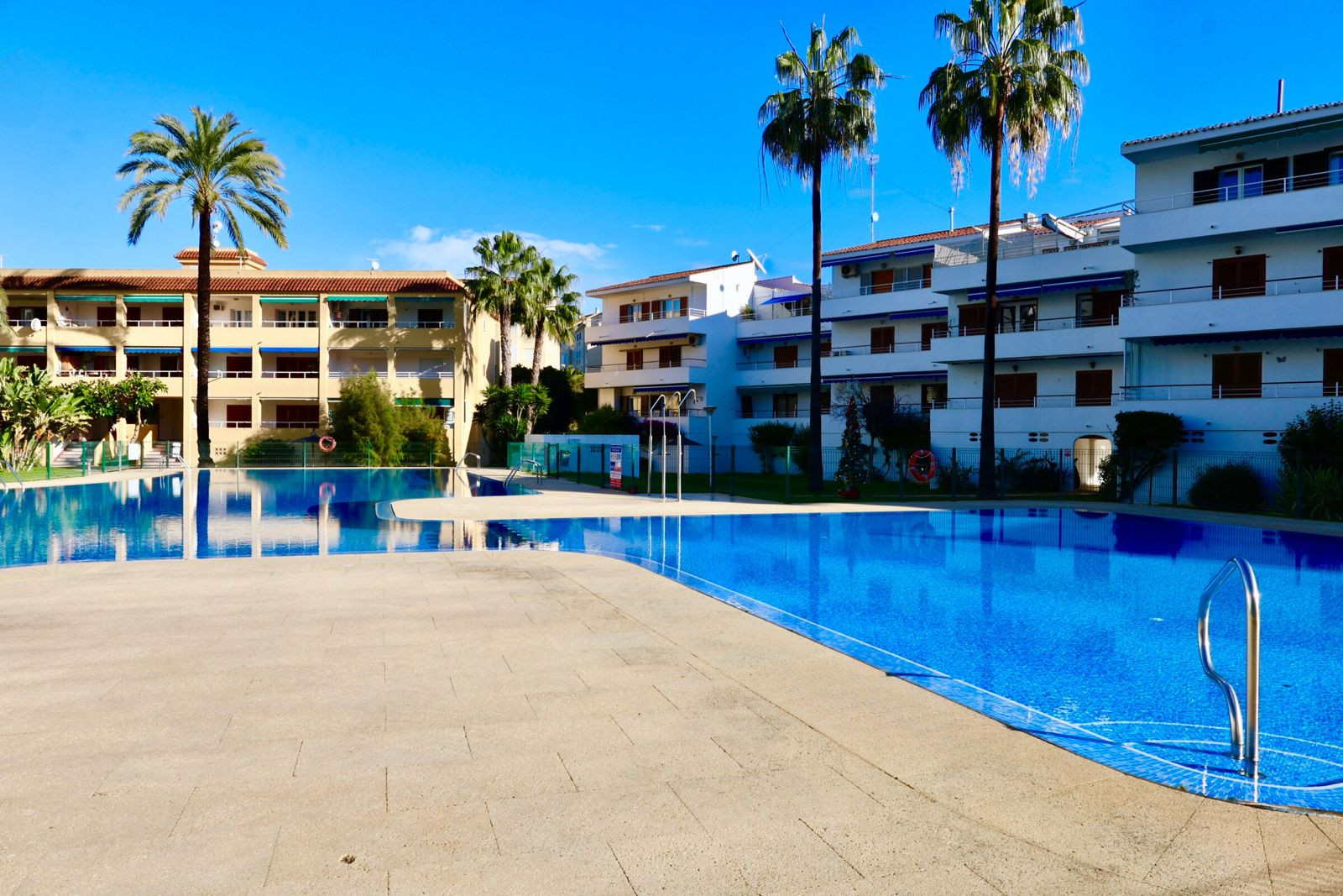 Großer Gemeinschaftspool mit blauen Mosaikfliesen, umgeben von einer hell gefliesten Terrasse und Palmen. Apartmentgebäude mit Balkonen und blauen Markisen unter klarem Himmel im Hintergrund. Großer Gemeinschaftspool mit blauen Mosaikfliesen, umgeben von einer hell gefliesten Terrasse und Palmen. Apartmentgebäude mit Balkonen und blauen Markisen unter klarem Himmel im Hintergrund.