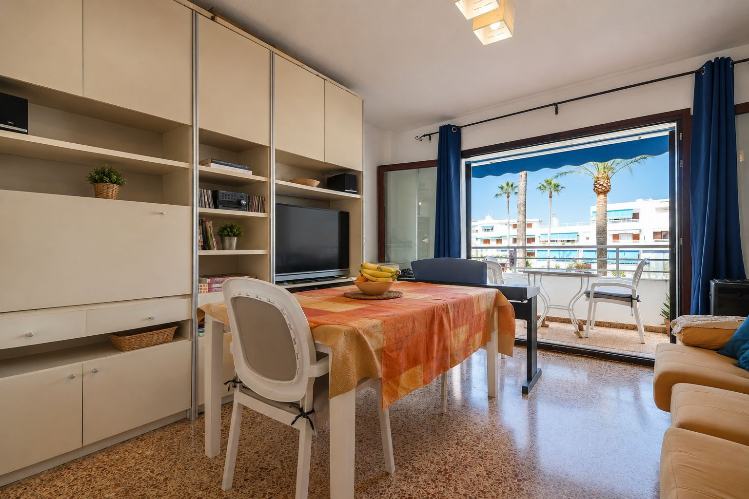 Heller Wohn-Essbereich mit Esstisch und integrierter Medieneinheit, der sich zu einer Terrasse mit Palmenblick und weißen Gebäuden öffnet. Heller Wohn-Essbereich mit Esstisch und integrierter Medieneinheit, der sich zu einer Terrasse mit Palmenblick und weißen Gebäuden öffnet.