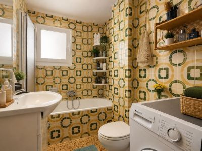 Das Badezimmer zeigt charakteristische gelb-grüne geometrische Fliesen, einen Waschtisch mit Unterschrank, eine Badewanne mit Duschwand und eine integrierte Waschmaschine, beleuchtet durch ein Fenster.