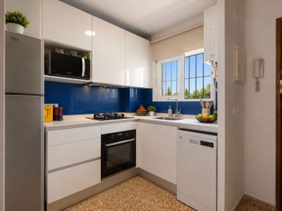 Moderne und funktionale Küche mit glänzenden weißen Schränken, heller Arbeitsplatte und blauem Spritzschutz. Ausgestattet mit integrierten Geräten und Fensterblick ins Grüne.